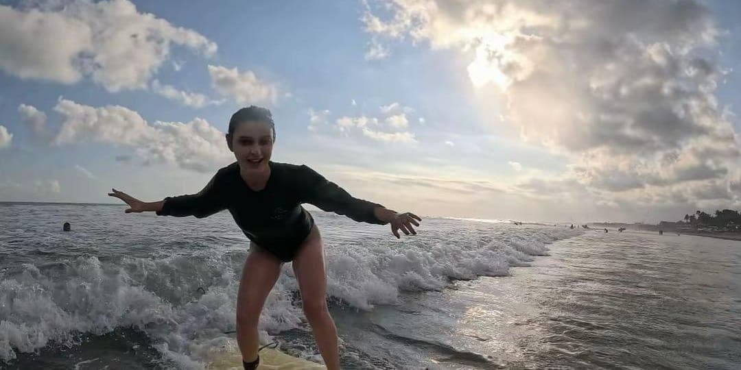 An older student riding a wave with her arms wide, under a broken sky at the end of the day