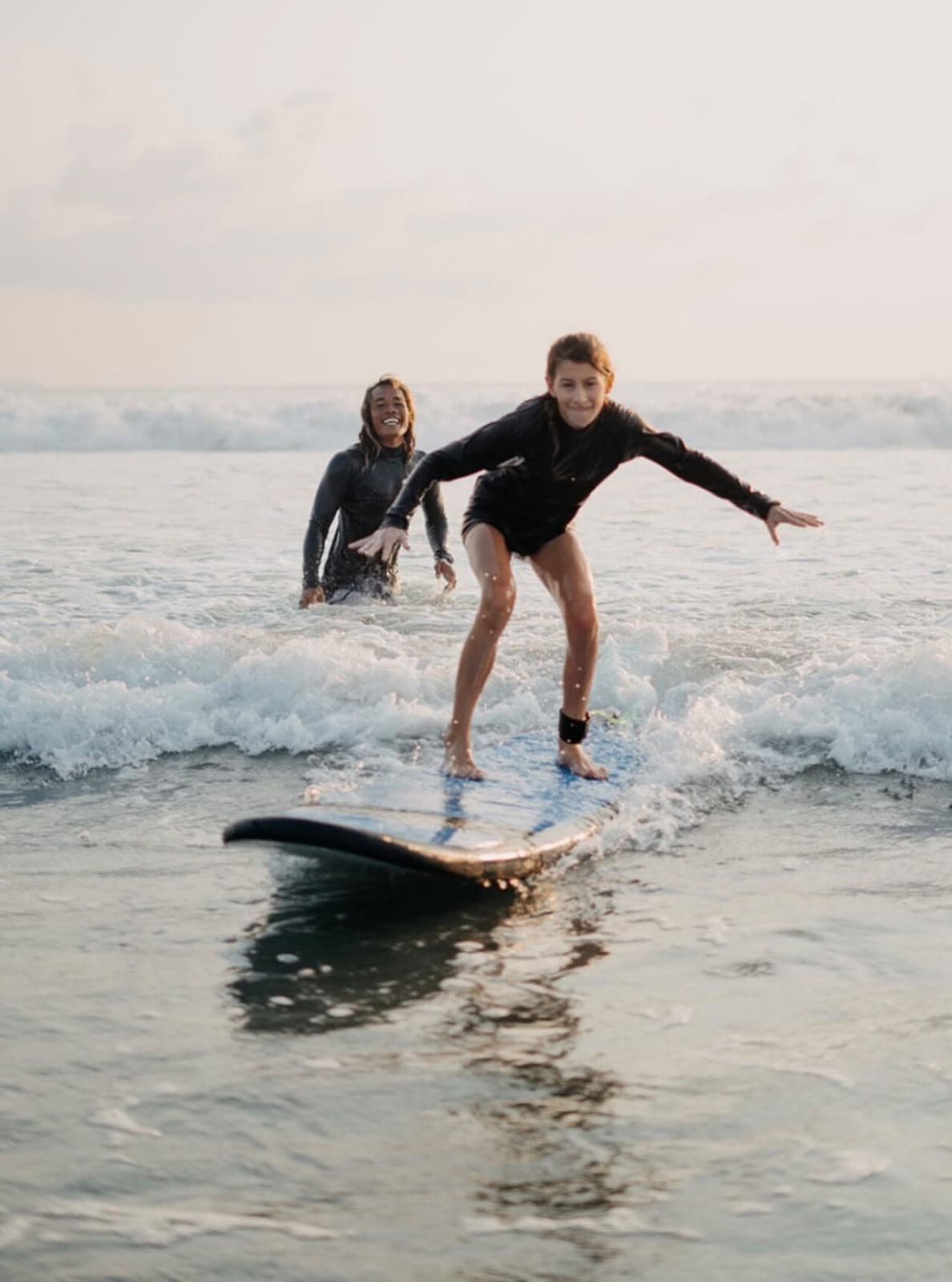 A student in a black wetsuit riding a wave with arms spread wide, her instructor cheering from the water behind her at golden hour