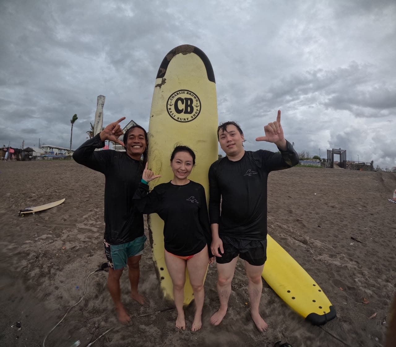 Three surfers throwing shakas beside a yellow board on an overcast beach, wearing Island Surf rash guards
